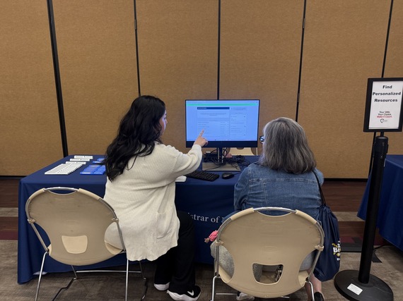 Two women sit at a table viewing a computer screen. One points at the screen while explaining. A sign nearby reads "Voter Registration Assistance.