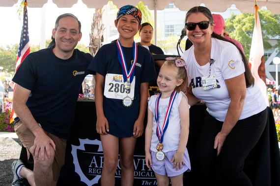 A smiling group of two adults and two children posing with medals in front of a podium at an outdoor event. The atmosphere is joyful and celebratory.