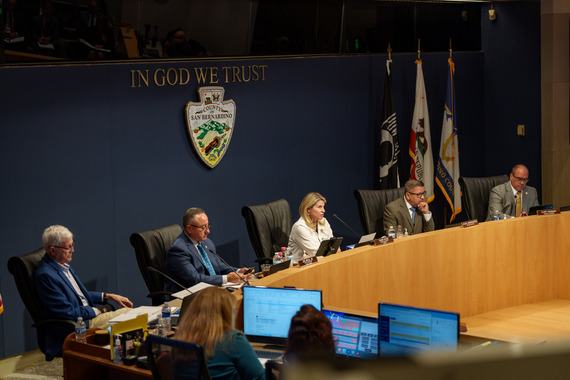 Officials seated at a dais beneath “In God We Trust” and the San Bernardino County seal.