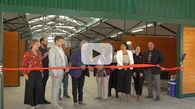 A line of people stand behind a red ribbon taped inside a horse barn while a woman in the center holds a large pair of scissors cutting the ribbon.