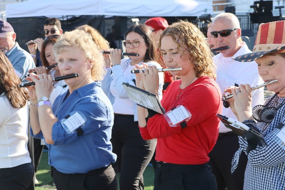 piccolo players perform together outdoors during a community symphony event, dressed in casual patriotic colors under bright afternoon sunlight.