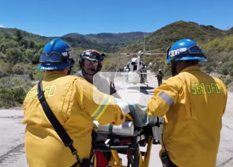 Three firemen in the national forest help a patient on a gurney to be airlifted to the hospital, with a video play button overlay indicating a video.