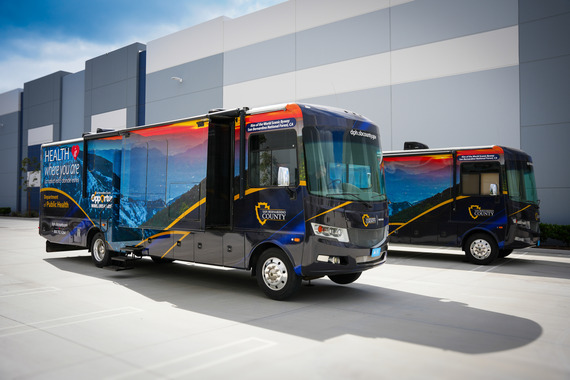 2 San Bernardino County Public Health mobile medical clinics parked outside a commercial building. The larger one shows mountain graphics.