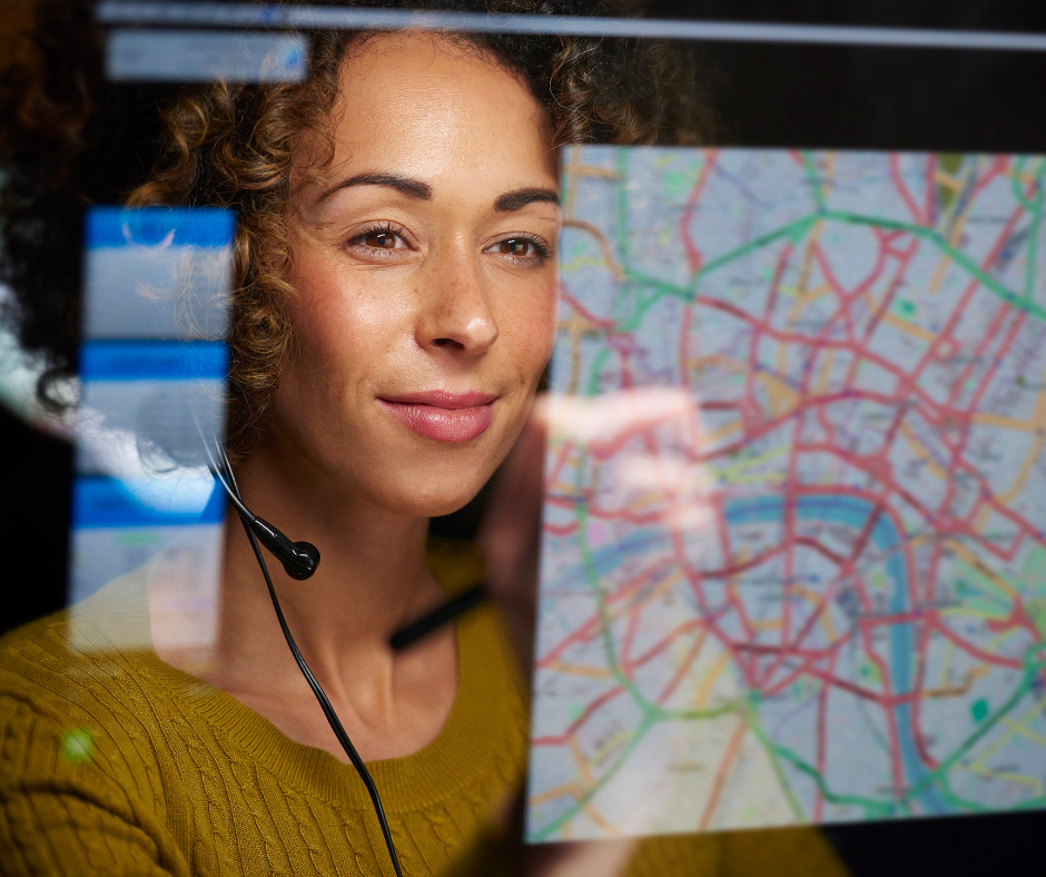 A curly haired woman in a gold sweater wears a phone operator headset while she stares pensively at a map on a transparent screen in front of her.