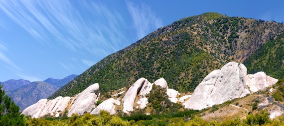 Large rock formations are in the foreground, and the background features a hillside covered with grass under a clear blue sky with wispy clouds.