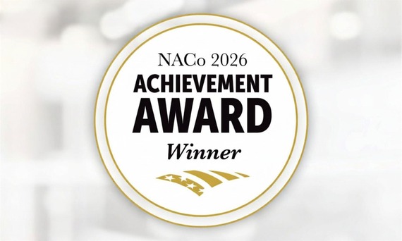 Circular gold-and-white seal with the text “NACo 2026 Achievement Award Winner” against a soft blurred background.