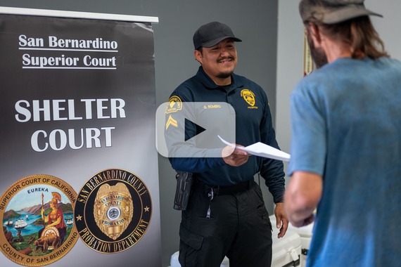 A Probation staff member hands a document to a man. Also visible is a San Bernardino Superior Court Shelter Court sign.