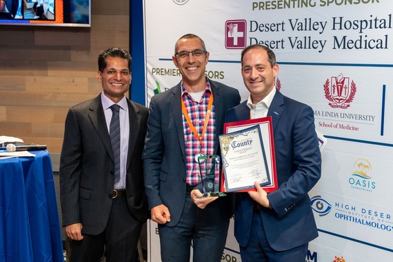 Three individuals are standing together. One person is holding an award and certificate. The backdrop displays logos for sponsors.