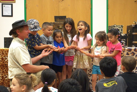 A group of excited children and a man hold a large snake during an educational event. The atmosphere is lively, with kids showing curiosity and joy.
