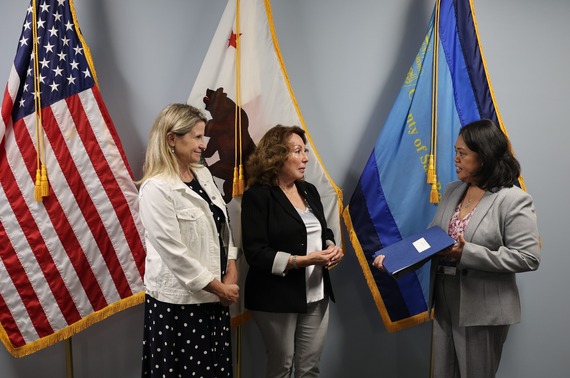 Three individuals are standing in front of some flags. They are engaged in conversation, with one holding a folder.