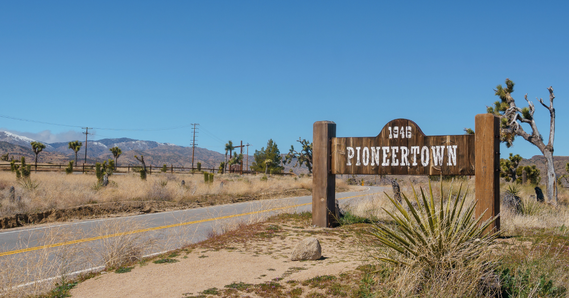 A wooden sign reading “1946 Pioneertown” stands beside a rural desert road with dry vegetation