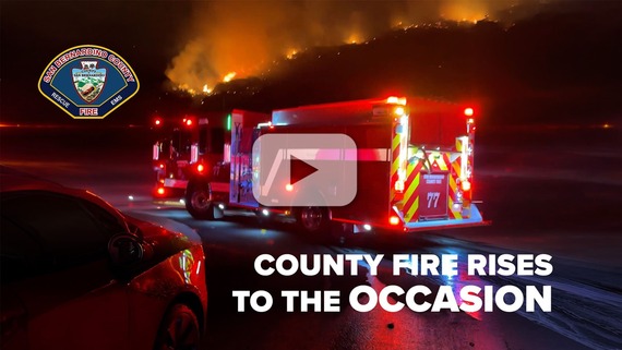 A nighttime scene shows a fire truck positioned on a road, responding to a significant fire on a hillside.