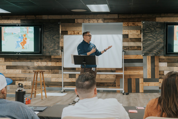 A person stands and presents to a group in a room with wooden wall panels.