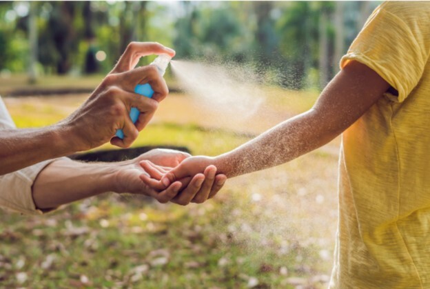 An adult sprays insect repellent onto a child’s arm outdoors in a sunny, grassy area.