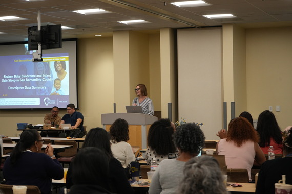 A person is speaking at a podium in a conference room, with an audience seated at tables.