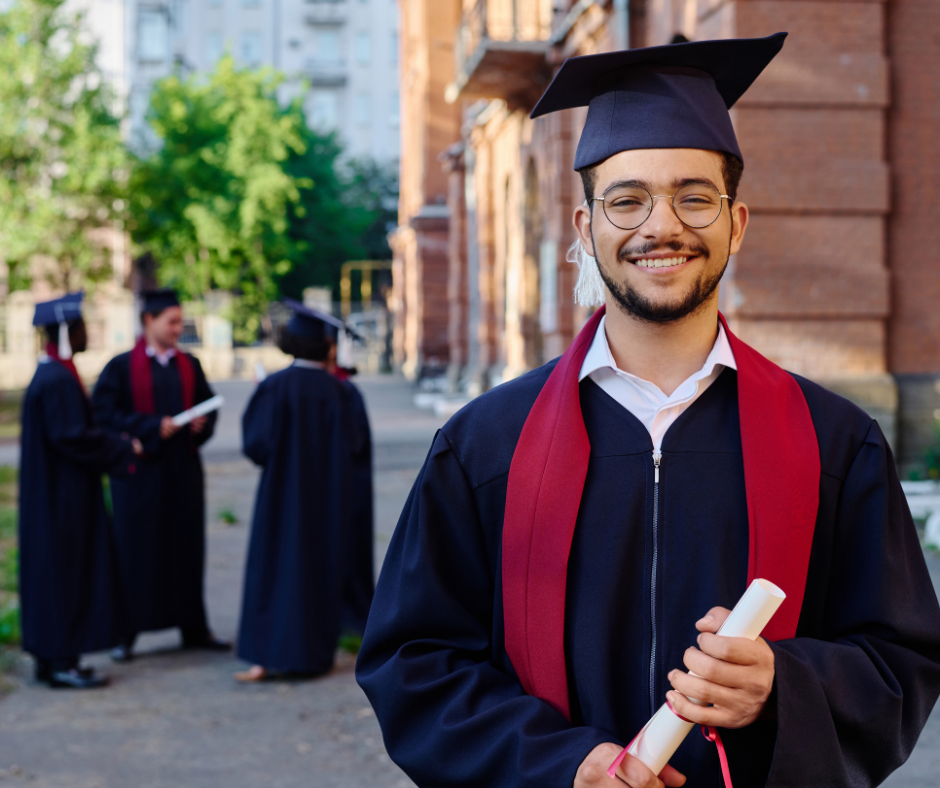 Smiling graduate in a black cap & gown with a red stole stands outdoors holding a rolled diploma with several other graduates in the background