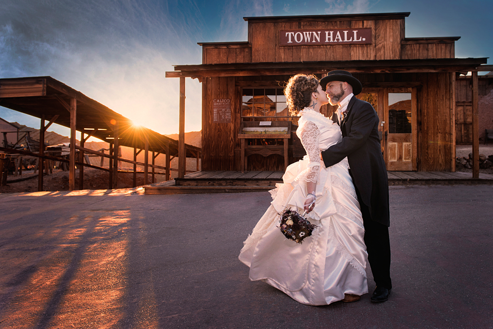 A bride and groom wearing traditional wedding clothing share a kiss at sunset at Calico Ghost Town in Yermo, California.