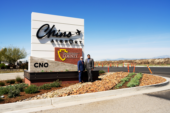 Two men stand beside the Chino Airport sign featuring the County logo and CNO code, with landscaping, mountains and a clear blue sky behind them.