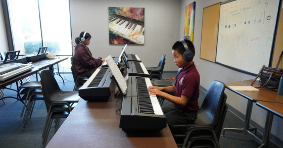 A young boy and a girl sitting in front of two piano keyboards with headphones on. 