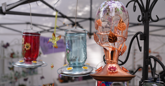 A close-up of three ornate glass hummingbird feeders hanging from a metal structure at an outdoor market. 