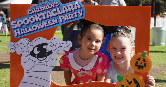 Two young children in Halloween costumes smile through an orange photo frame decorated with a cartoon mummy, jack-o’-lanterns