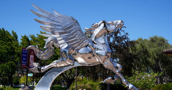 Large, polished metal sculpture of a Pegasus with outstretched wings appears to leap into the air against a clear blue sky. 