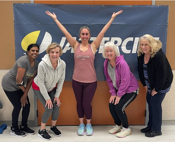 "Five women smiling in front of a Jazzercise sign. The middle woman has her arms up, and the others are bending forward."