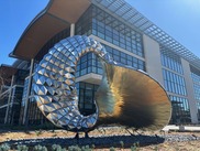 Reflective triangular sculpture in front of a modern glass building.