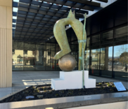 Modern sculpture of an abstract figure over a sphere near a building with glass windows.