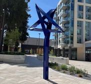 Blue abstract geometric sculpture in an urban plaza with a modern building in the background.