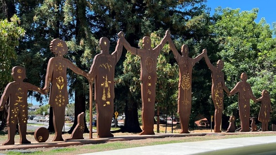 A row of metal silhouettes of people holding hands, with cut-out designs, set outdoors against a backdrop of trees and blue sky.