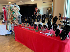 "Handcrafted jewelry and holiday accessories displayed on tables with red tablecloths at a boutique event."