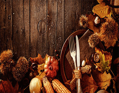 Rustic autumn table setting with a fork and knife on a brown napkin surrounded by leaves, corn, chestnuts, and small gourds on a wooden surface.