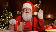 Santa Claus smiling and waving while sitting at a festive table with a wrapped gift, Christmas tree, and warm holiday decorations in the background.