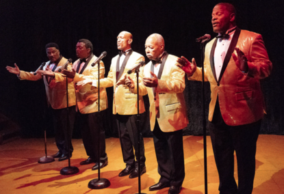 "Five men wearing gold blazers and black pants sing on stage into microphones, performing together under warm lighting with a dark curtain backdrop."