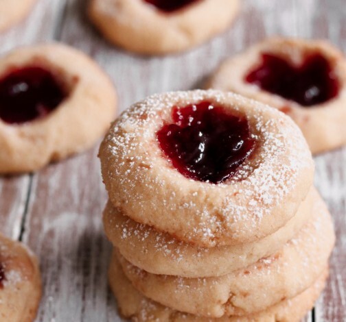 "Stack of thumbprint cookies filled with red jam and dusted with powdered sugar on a rustic wooden surface."