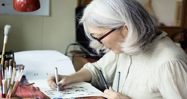 “Older woman with gray hair and glasses drawing at a desk, surrounded by art supplies.”