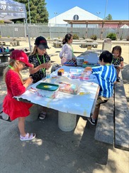 Children work together at an outdoor picnic table creating bead crafts and drawings during a supervised recreation activity.