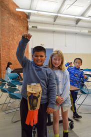 Two children smile indoors while holding a handmade paper craft, with other children and chairs visible in the background during a recreation activity