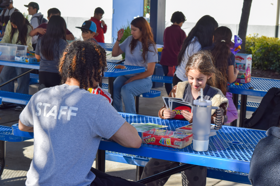Children sit at outdoor tables playing board games with a recreation staff member during a supervised program at a park.