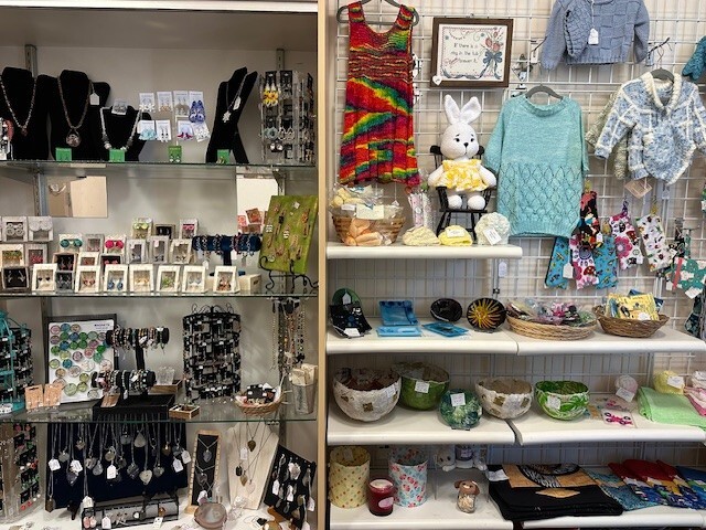 Craft fair display shelves show handmade jewelry, knitted clothing, stuffed toys and small gifts neatly arranged for sale.