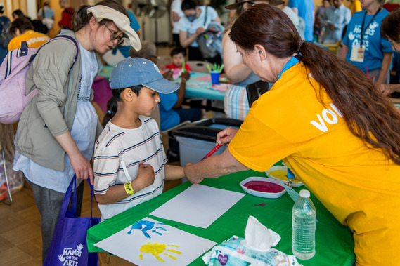 Volunteer helps a child make colorful handprint art at a busy indoor community arts activity table.