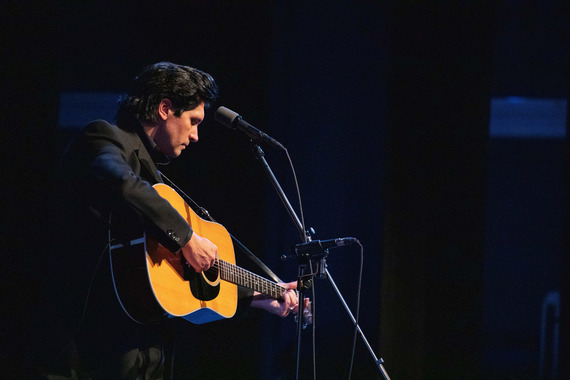 Musician plays an acoustic guitar onstage beside a microphone.