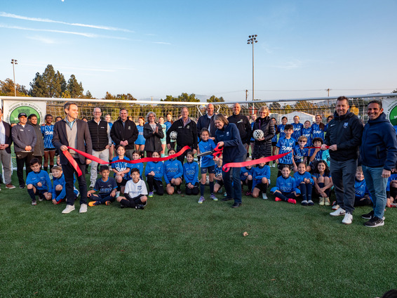 An image of Cindy Darling cutting a red ribbon on a soccer field with other soccer players, staff, and City employees around.
