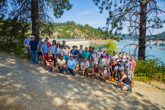 A group of people in front of a large reservoir and dam. 