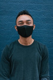 An individual stands against a wall with a mask on