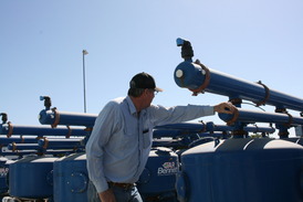 A man in a cap and striped shirt inspects large blue farming equipment under a clear sky.