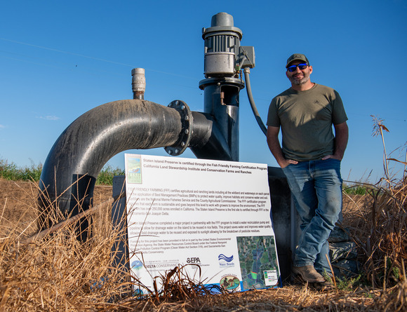 Farmer standing in front of a water pump that has a sign leaning against it