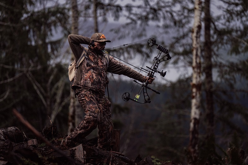 A big-game hunter dressed in camoflauge draws his bow in the forest.