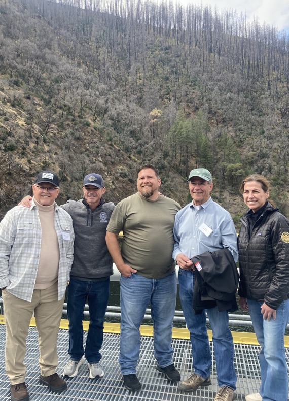 Five legislative members standing on dam with burn scarred river canyon in the background.